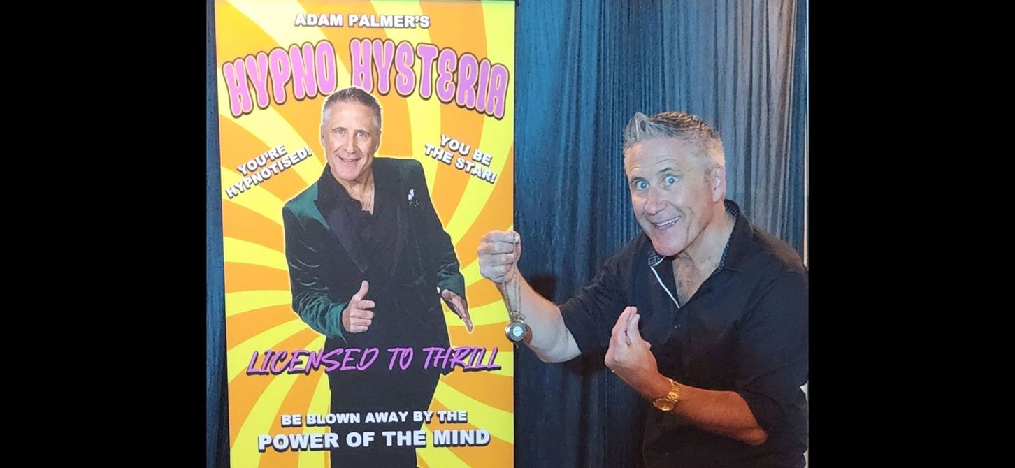 Man standing next to a poster of himself, smiling, pointing and gesturing with his hands.
