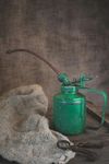 Groene oliekannetje met tuit, naast een jutezak en roestig gereedschap op een houten ondergrond.