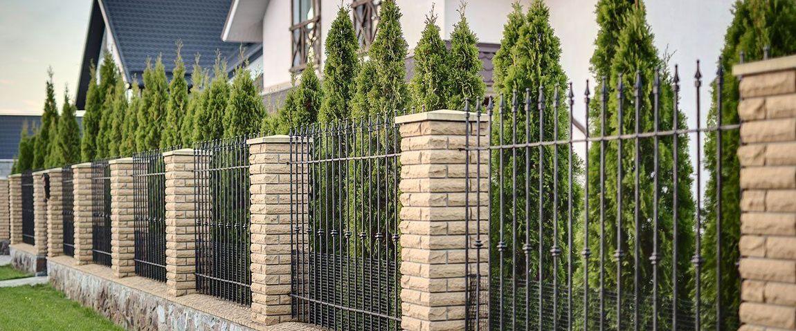 Fence with brick pillars, ironwork, and green trees in front of a house.