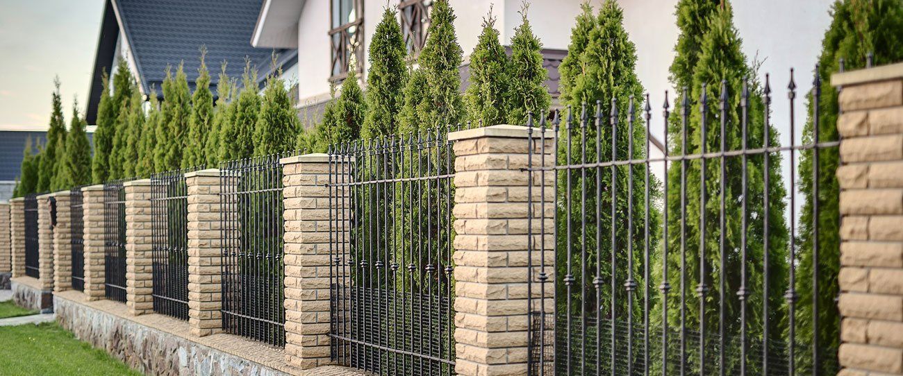 Fence with brick pillars, ironwork, and green trees in front of a house.