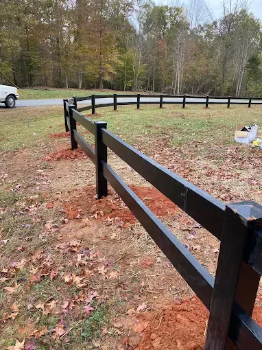 Black two-rail fence in a field with trees and a road in the background.