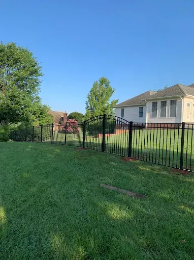 Black metal fence with arched gate surrounding a green lawn and white house.