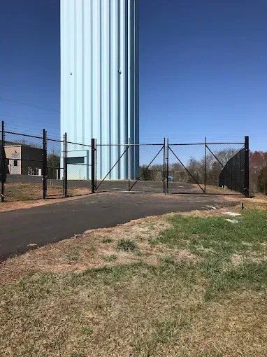 A tall blue water tower behind a black fenced gate, with a paved path in front, against a blue sky.