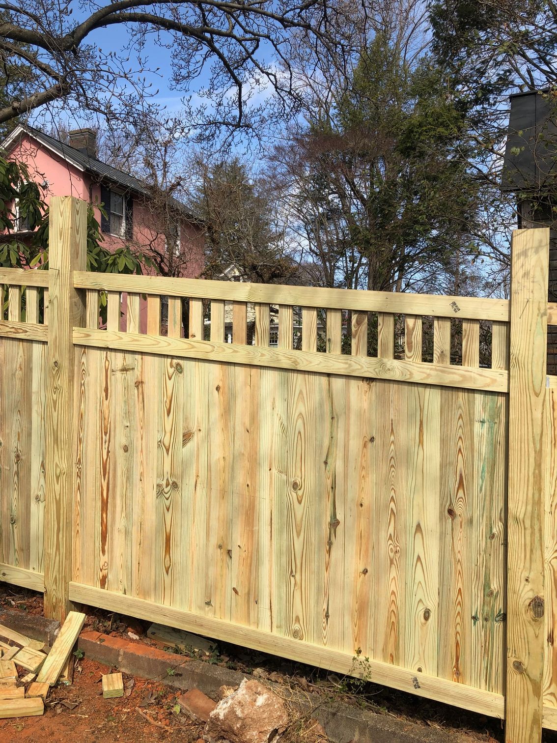 A wooden fence with vertical planks and a decorative top, in front of a house and trees.
