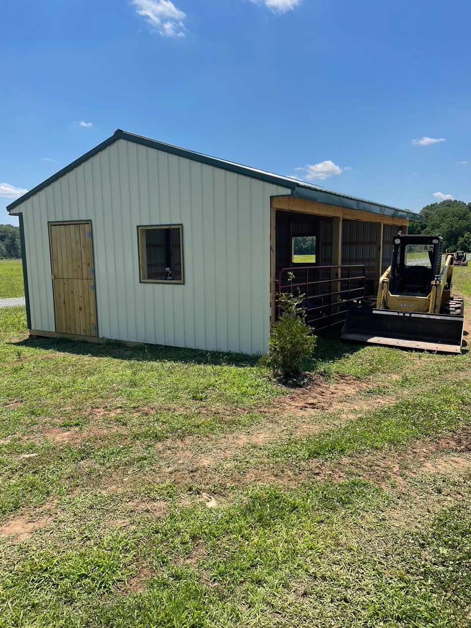 White shed with green roof and screened porch; yellow skid steer nearby in grassy field.