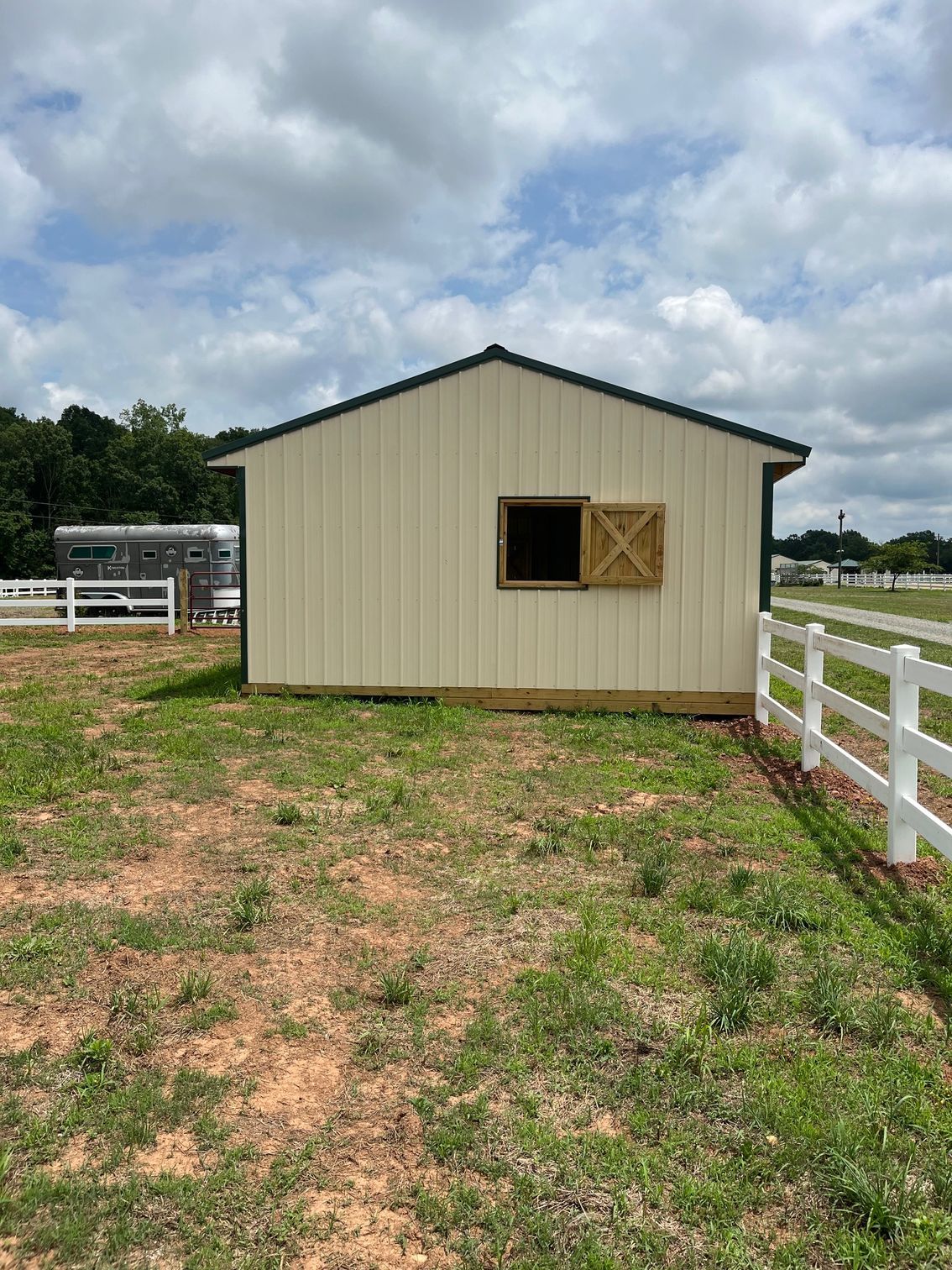 Small beige barn with a window, wooden shutters, and white fence in a grassy field.