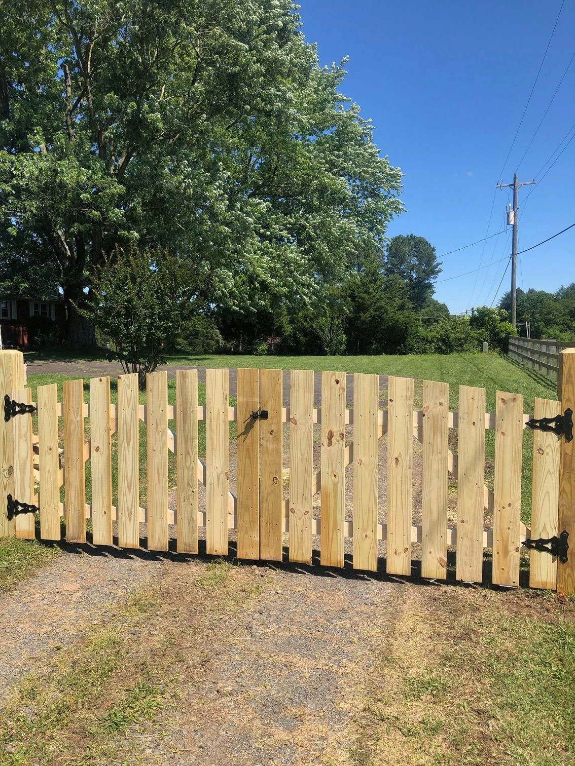 Wooden gate with vertical planks, black hinges, and a black handle. Opens to a green yard, blue sky.
