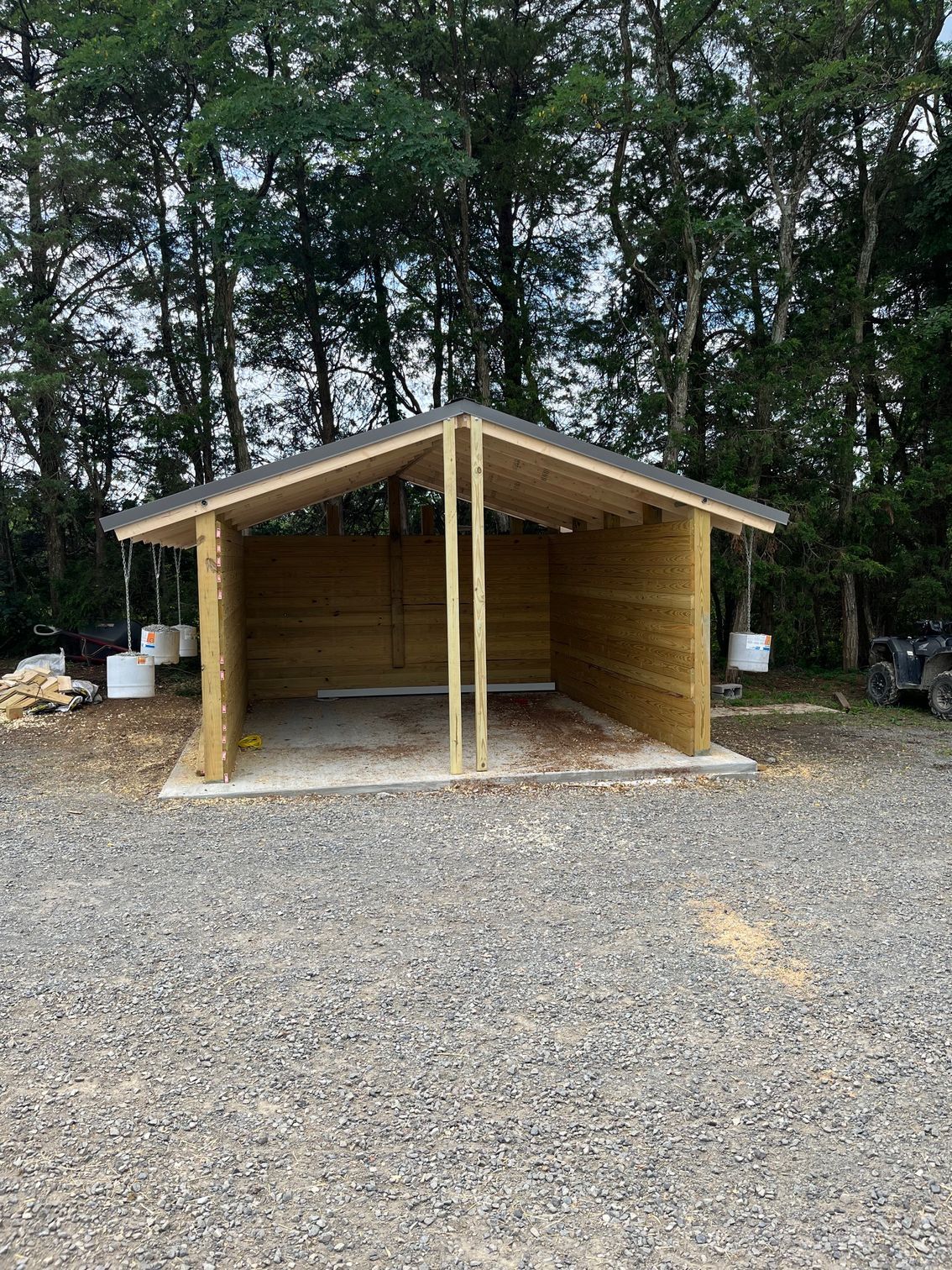 Wooden shed with open sides, two stalls, and a gray roof, set on gravel. Trees in the background.