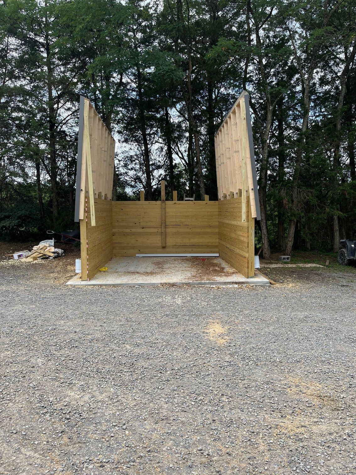 Wooden structure under construction, set on a gravel driveway with a backdrop of tall trees.
