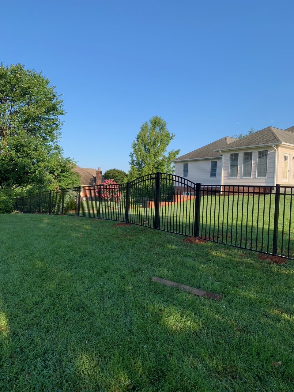 Black metal fence partially collapsed on green lawn, next to a house and trees under a blue sky.