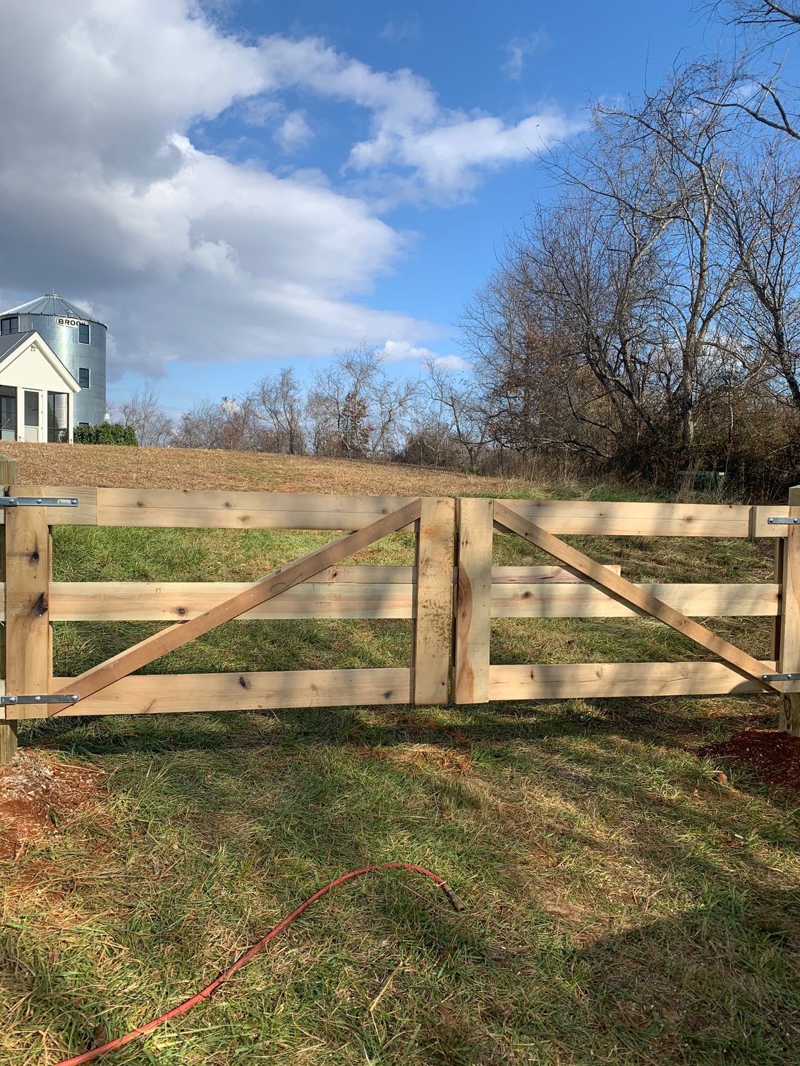 Wooden farm gate, light brown, with a diagonal brace. Grassy field, clear sky, trees.