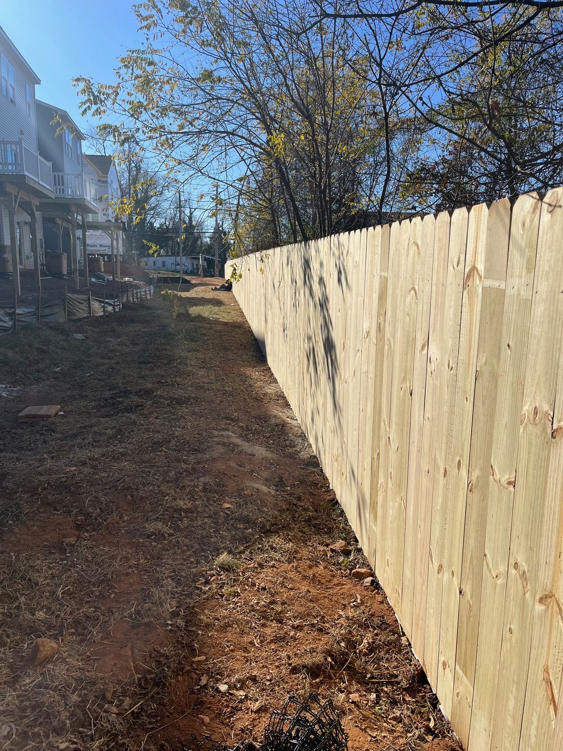 Wooden fence bordering a dirt path with a residential backdrop under a bright blue sky.