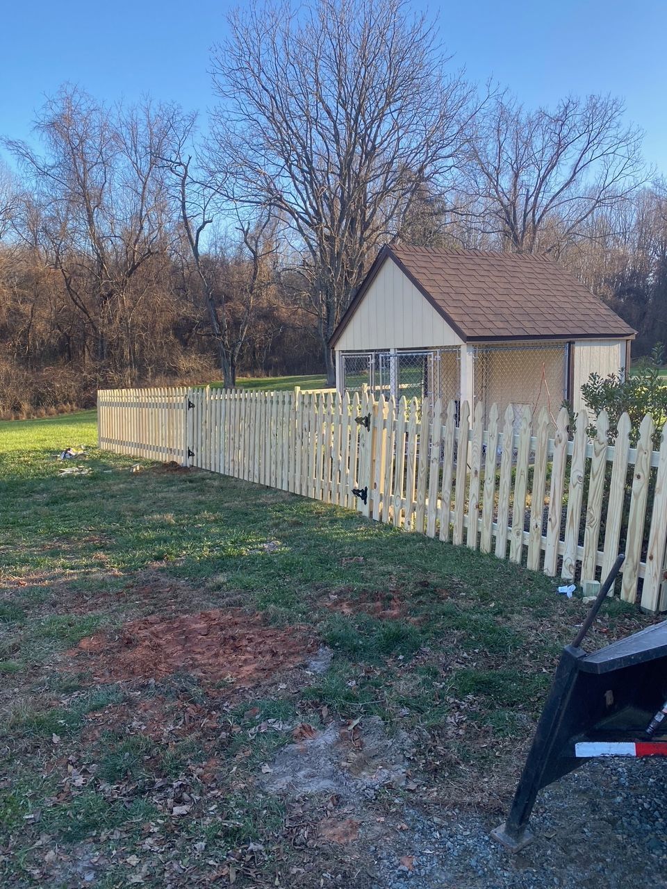 A light brown picket fence surrounds a small building, with a blue sky and trees in the background.