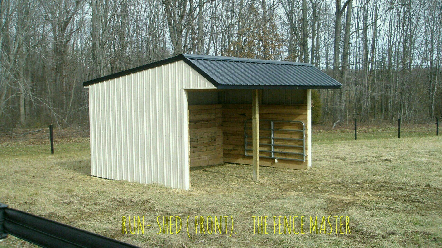A small, open-fronted beige and grey shelter in a field with trees in the background.