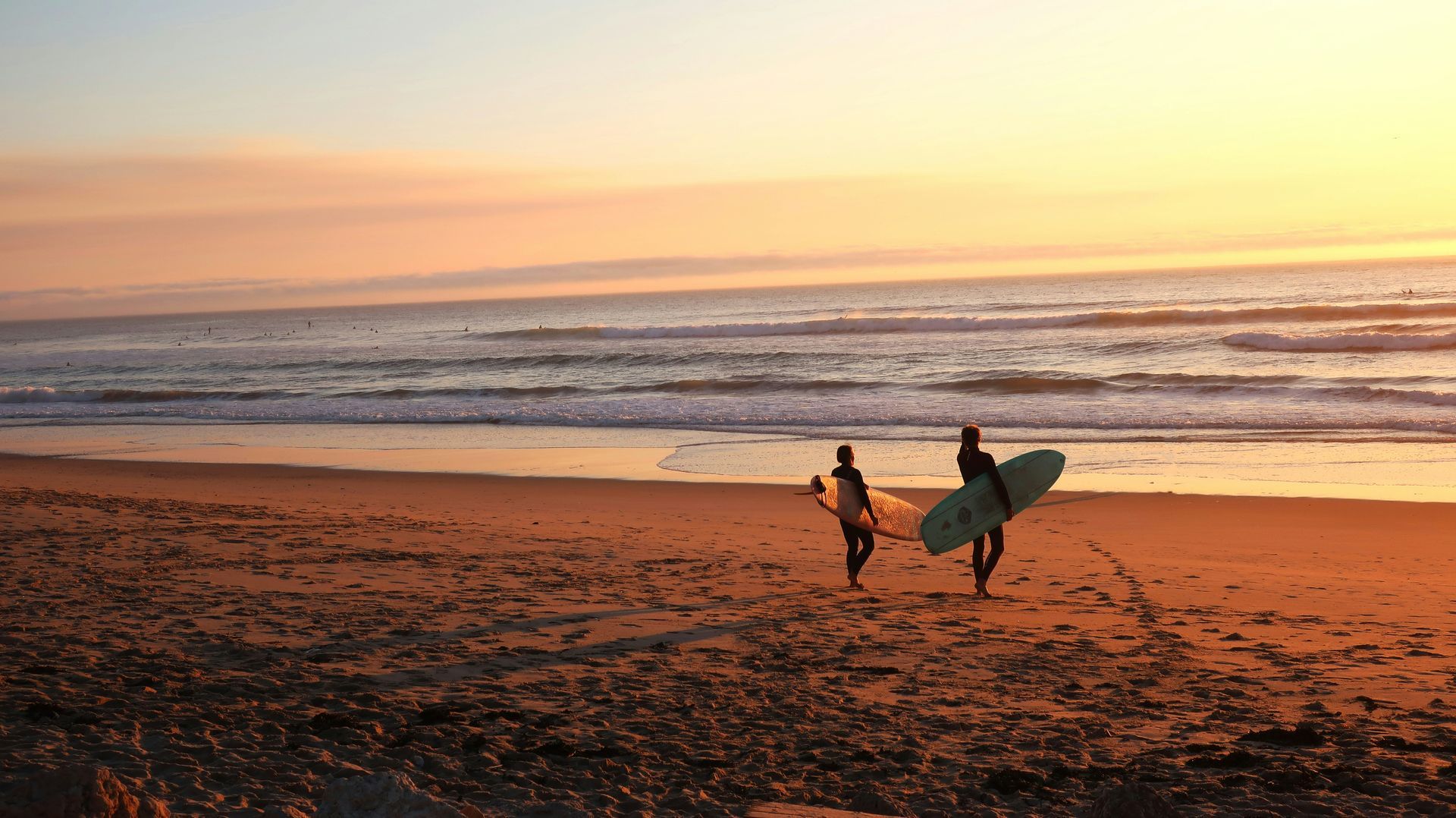 Two surfers walking with boards on a sandy beach at sunset, waves and orange sky in the background