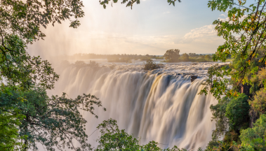 Victoria Falls with trees in the foreground and the sun shining through the trees.