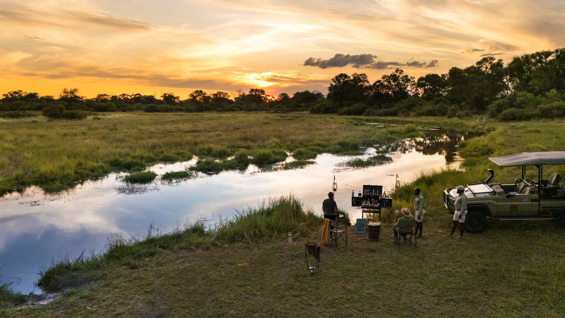 Sunset over a river with safari vehicle and chairs, two figures seated.