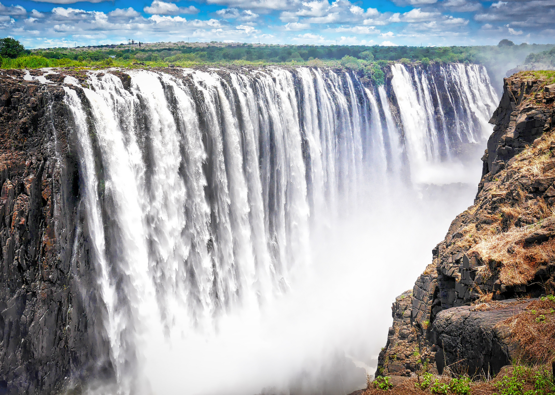 Victoria Falls cascading down a cliff face into a misty gorge, under a blue sky with scattered clouds.