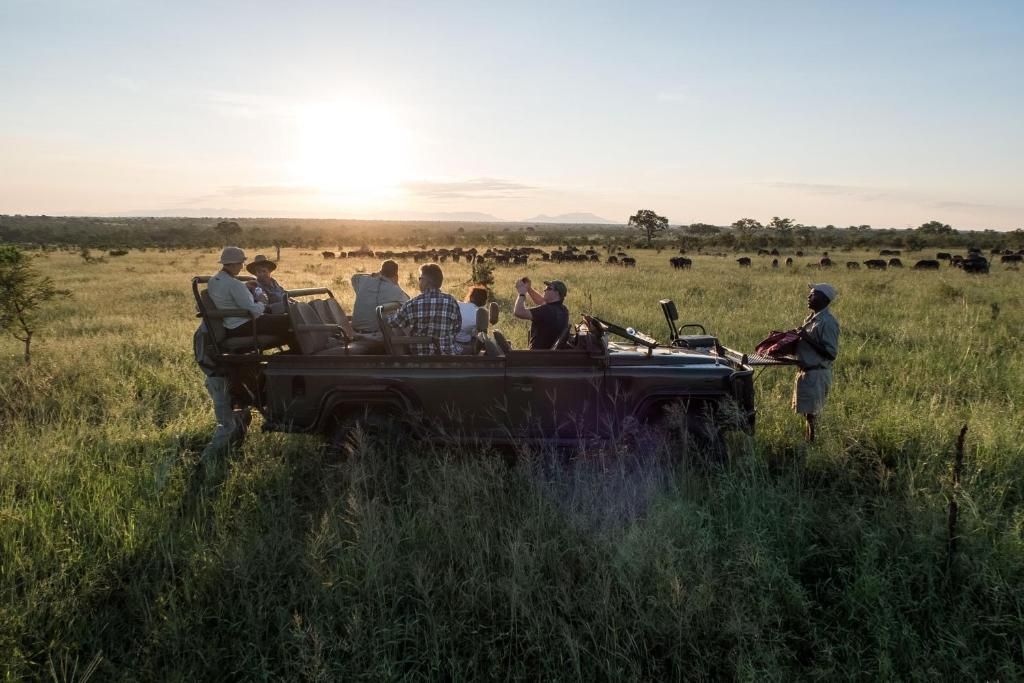 Safari vehicle in a grassy field with people, set against a sunset and herds of animals.