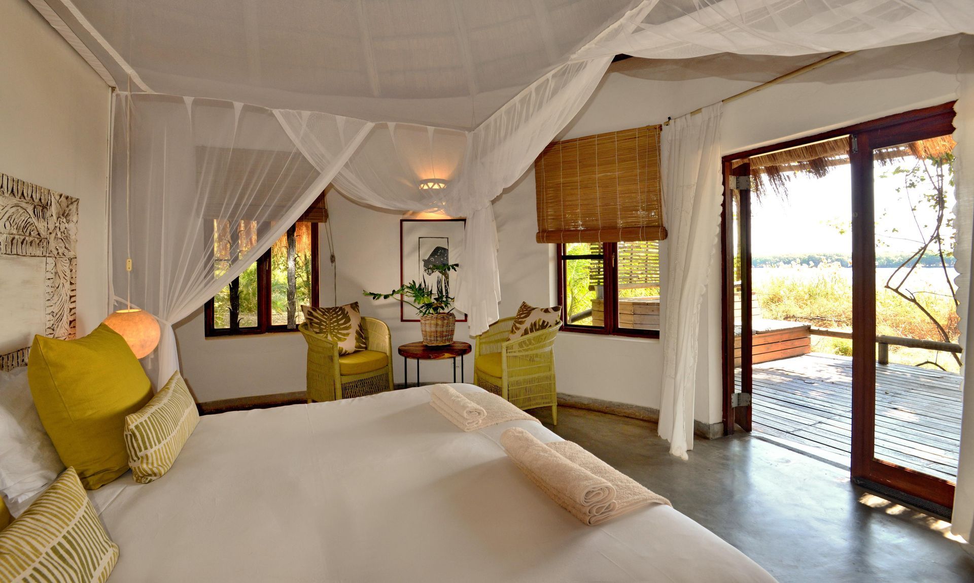 Bedroom with white canopy bed, open doors to balcony, and two yellow chairs. Chobe Bakwena Lodge
