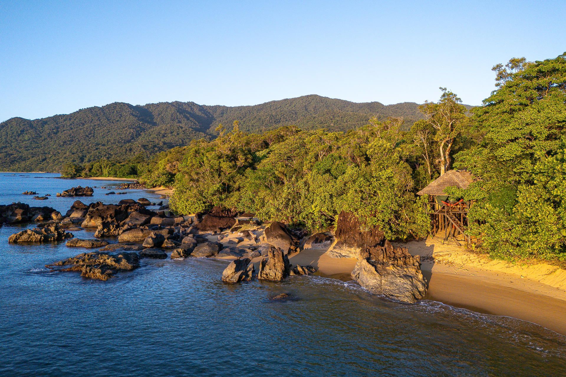 Beach with rocky shore, green trees, and hut, with a forested mountain in the background.