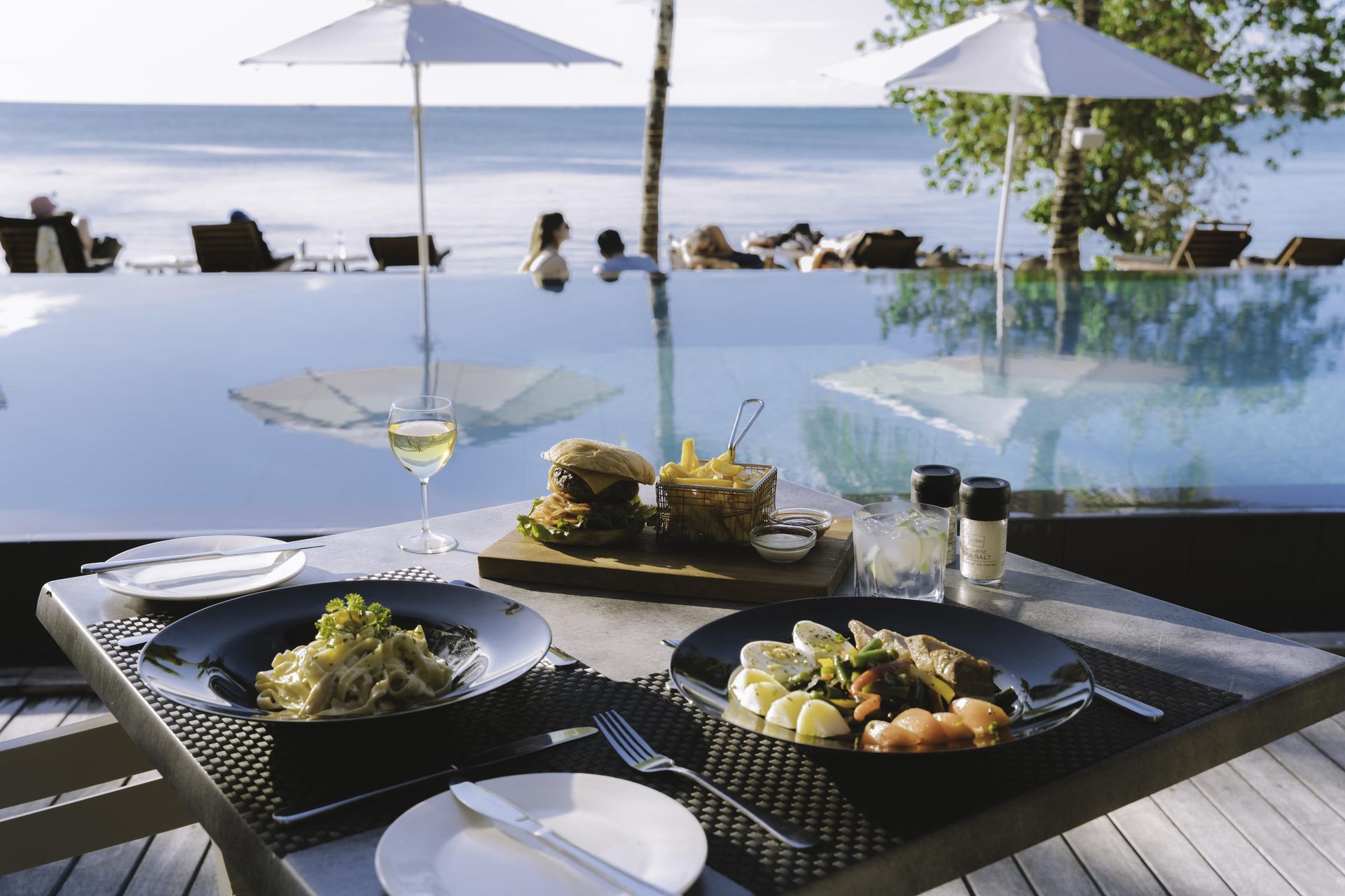 Table with food and drink, overlooking an infinity pool and ocean, with people relaxing in the background.