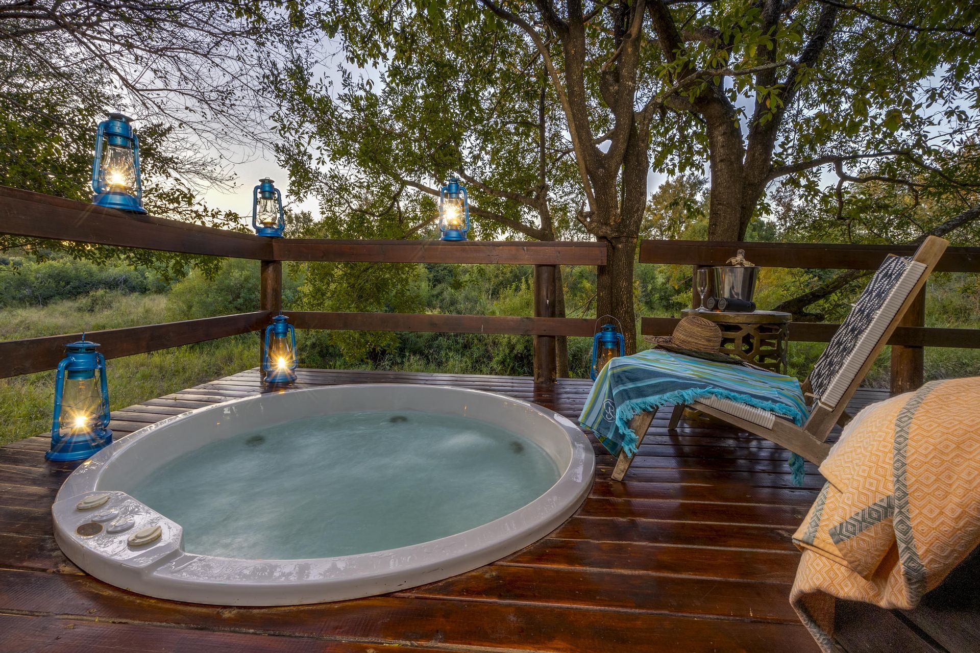A relaxing outdoor hot tub on a wooden deck, with lanterns, trees, and a lounge chair. Sabi Sabi LIttle Bush Lodge