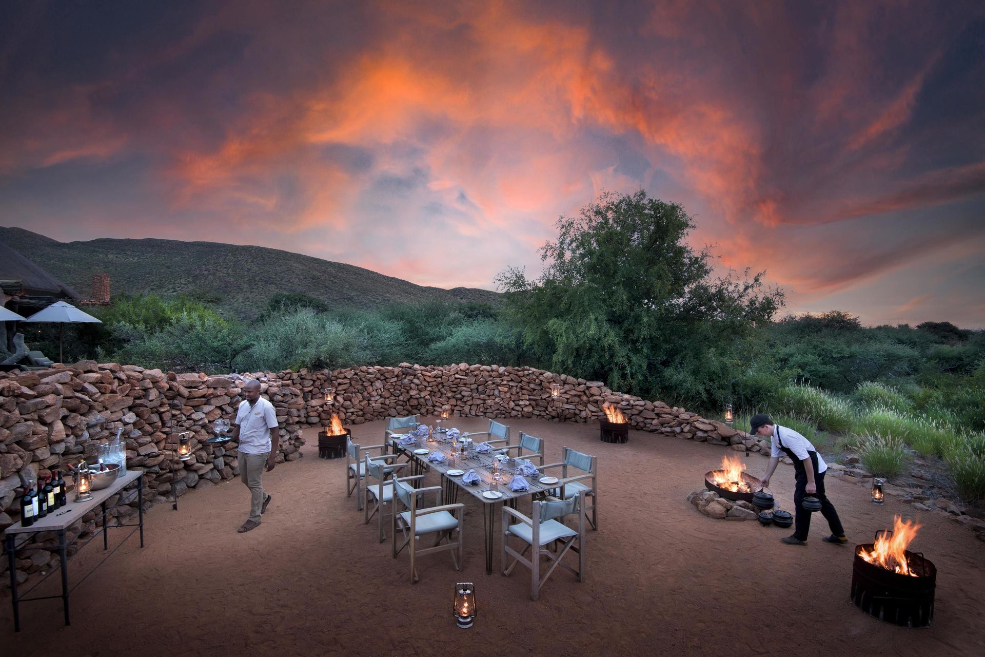 Outdoor dinner setting at sunset. Tables set, staff serving, fires burning. Lush landscape in background.