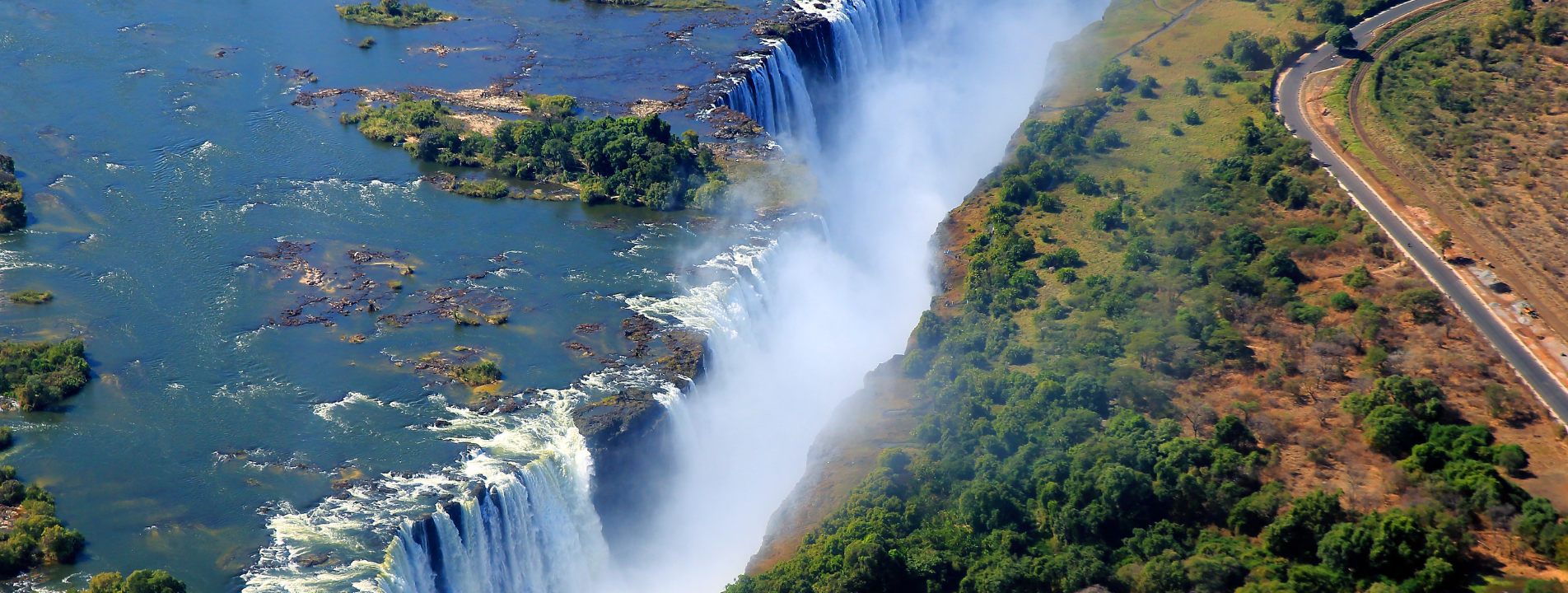 Aerial view of Victoria Falls in Zambia and Zimbabwe. The Zambezi River flows over the falls with surrounding lush green forest and a road.