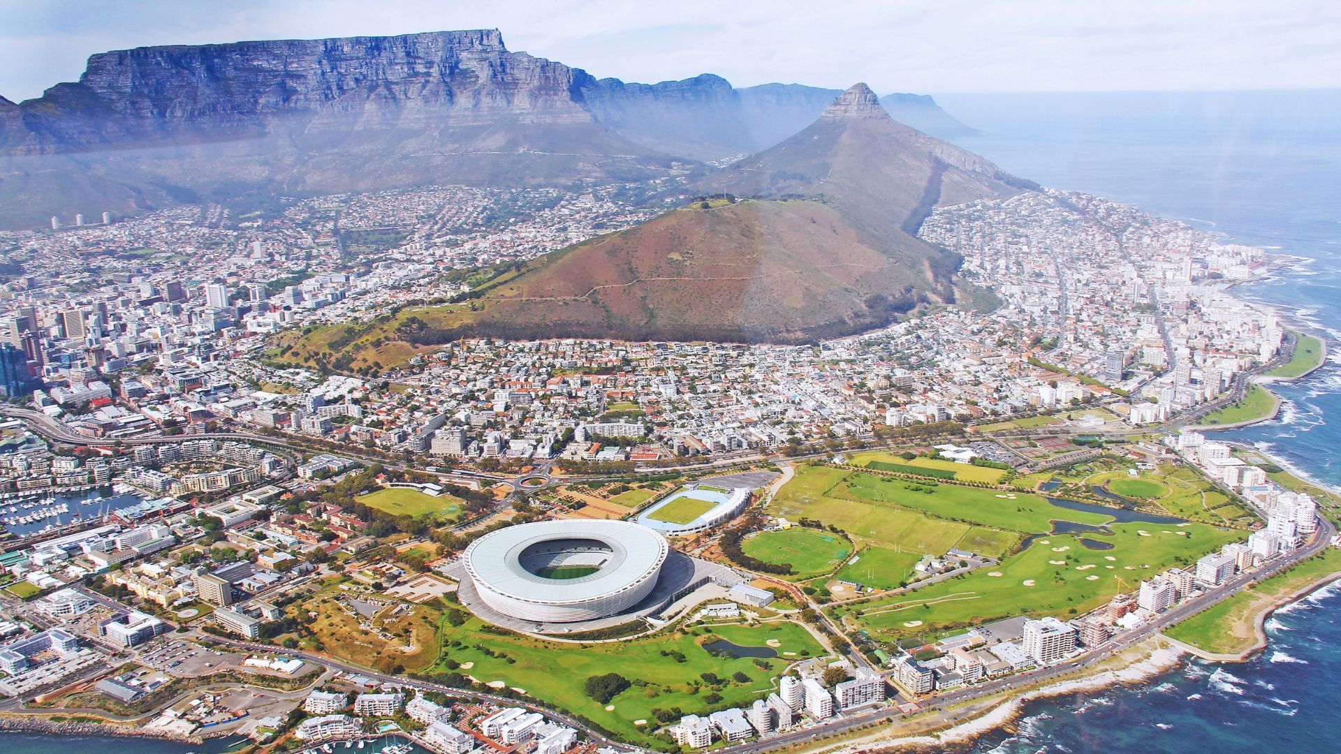 Aerial view of Cape Town, South Africa, with Table Mountain in the background and the Cape Town Stadium near the coastline.