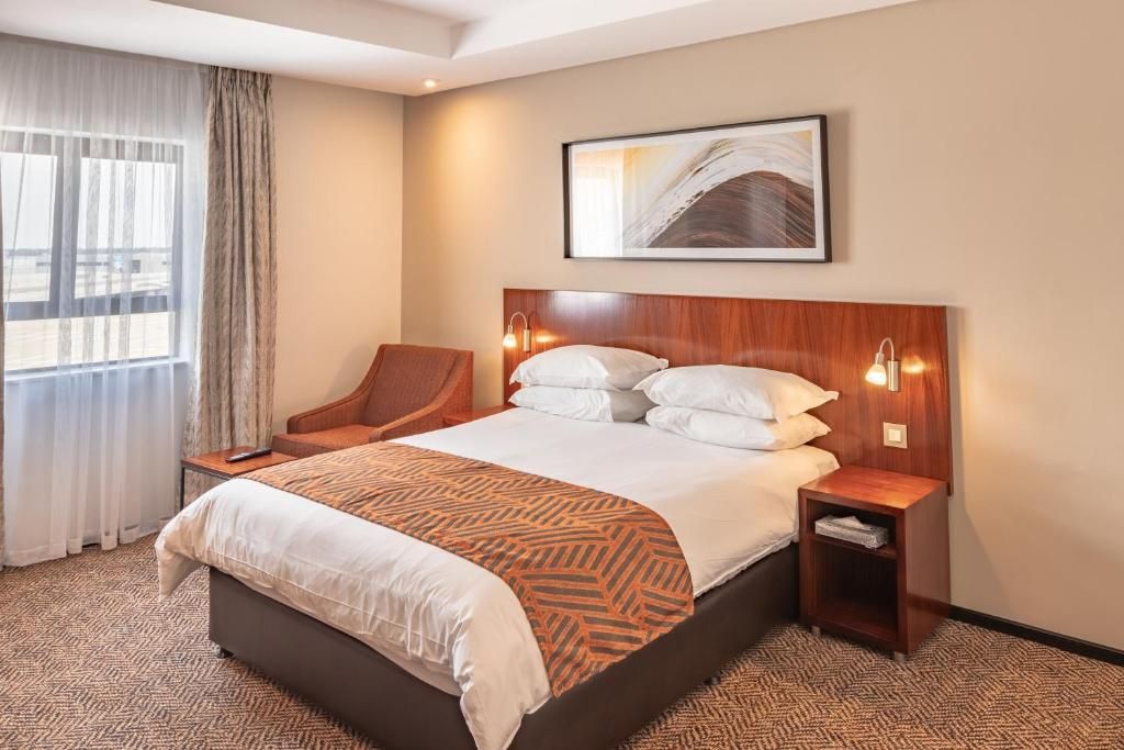 Hotel room with a double bed, armchair, nightstand, window, and abstract artwork.