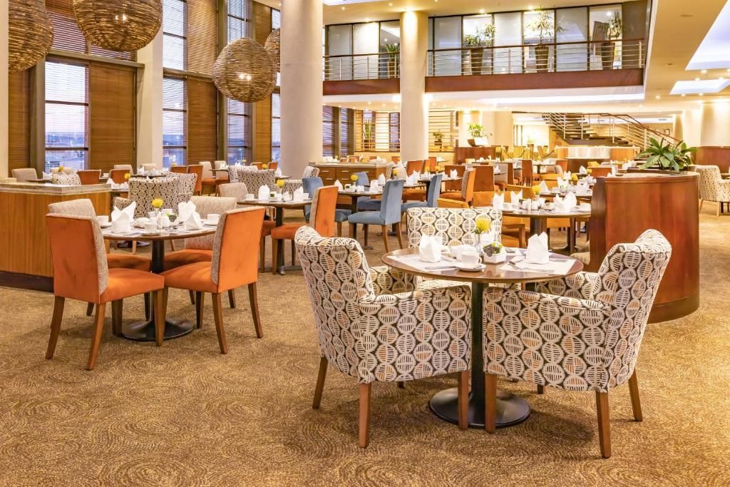Restaurant interior with tables, chairs, and large windows. Orange and patterned chairs, carpet flooring.