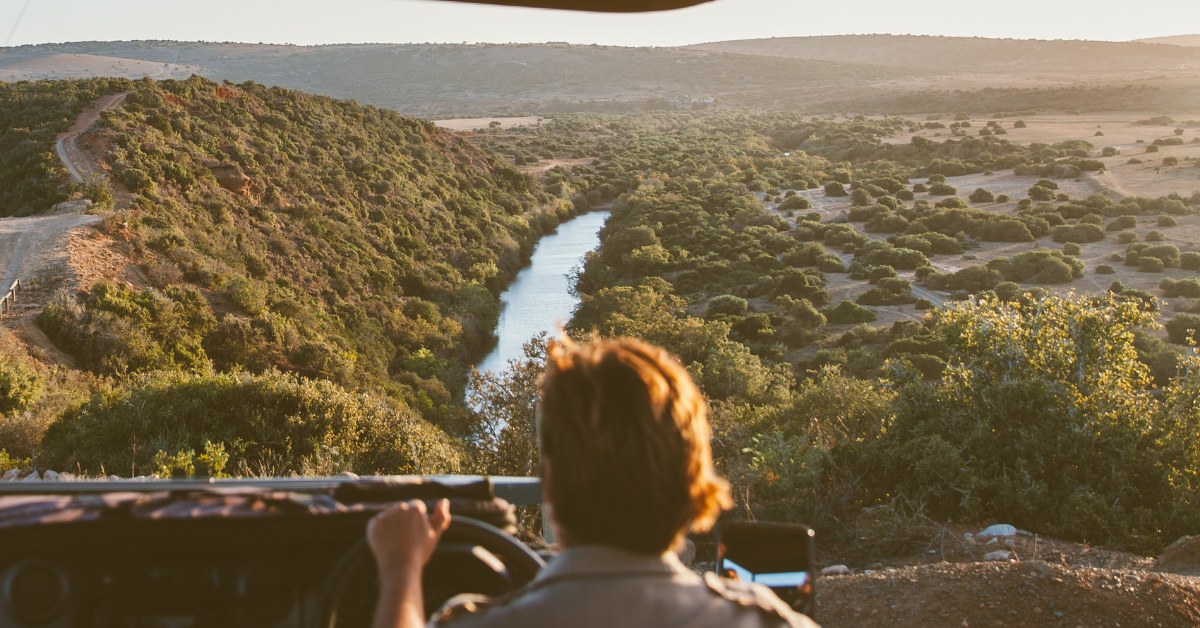 Person driving a jeep, looking at a river valley with a dark, unidentified object in the sky.