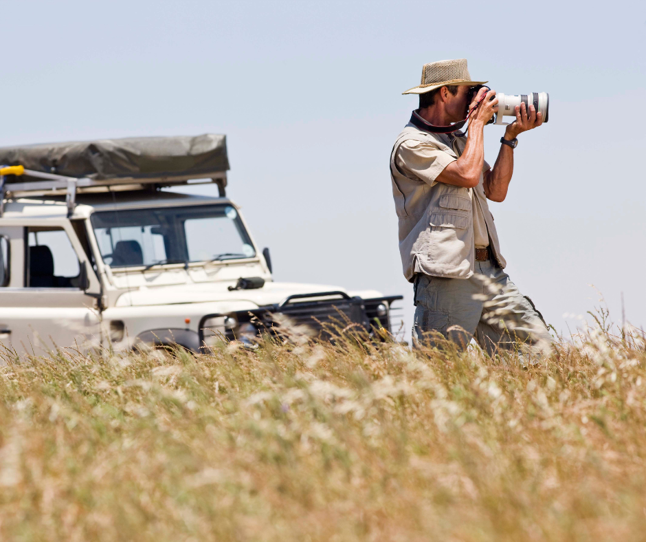 Man in safari attire photographs with a long lens, white jeep in background, tall grass, sunny day.
