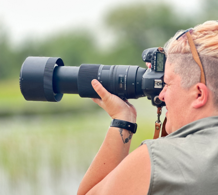 Photographer with a large lens, partially shaved head, outdoors by water, taking a photo.