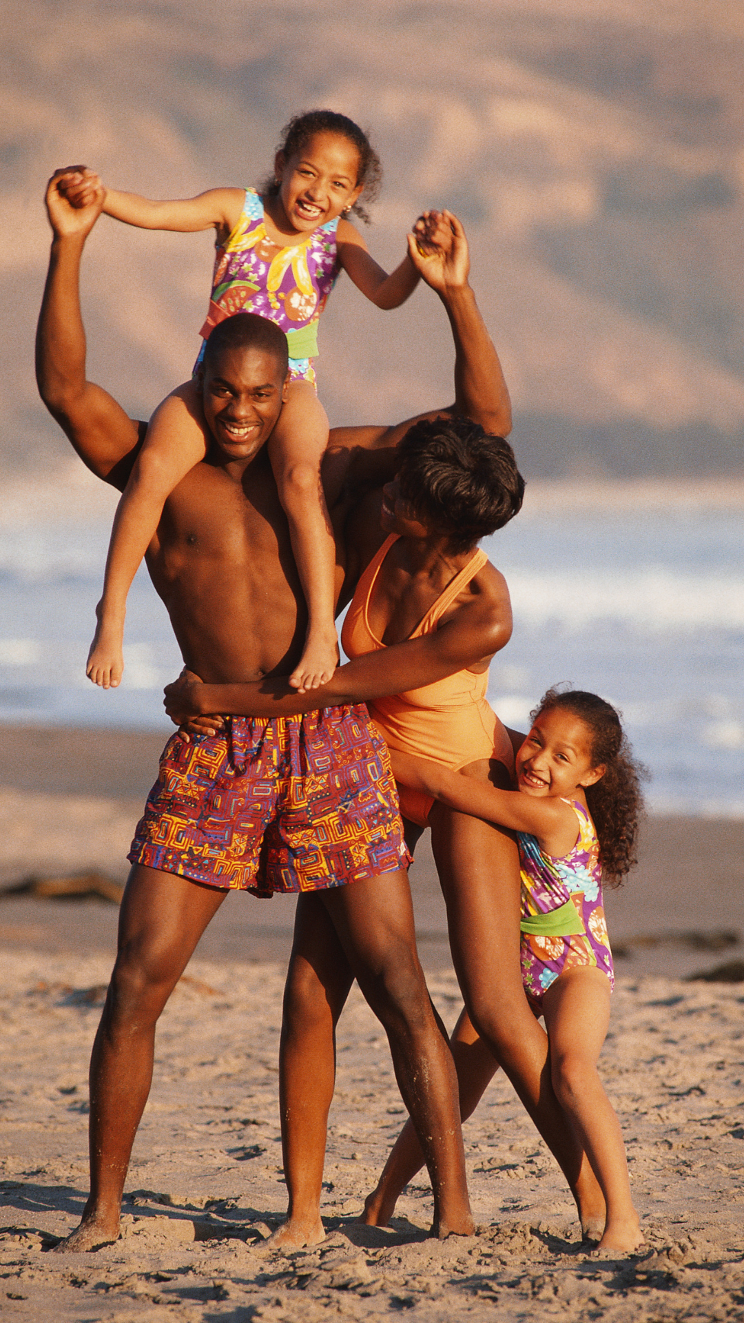 Family of four smiling on the beach, father holding daughter on shoulders, other daughter hugging mother.