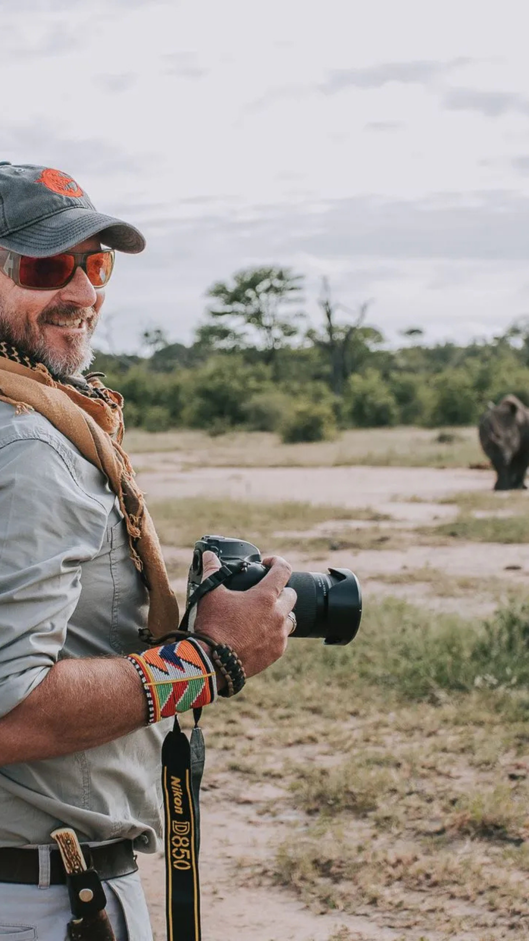 Photographer smiling, holding camera, in safari setting; elephant in background.