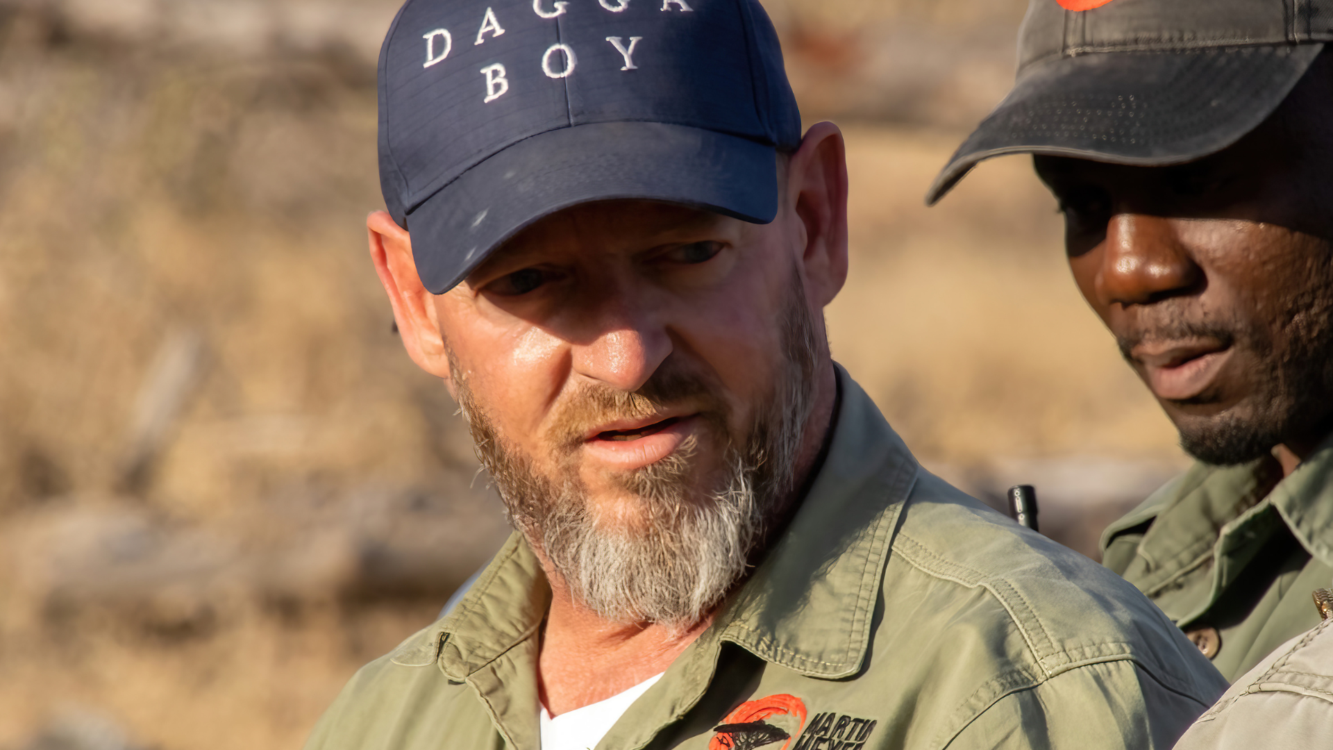 A man with a beard wearing a blue hat is standing next to another man.