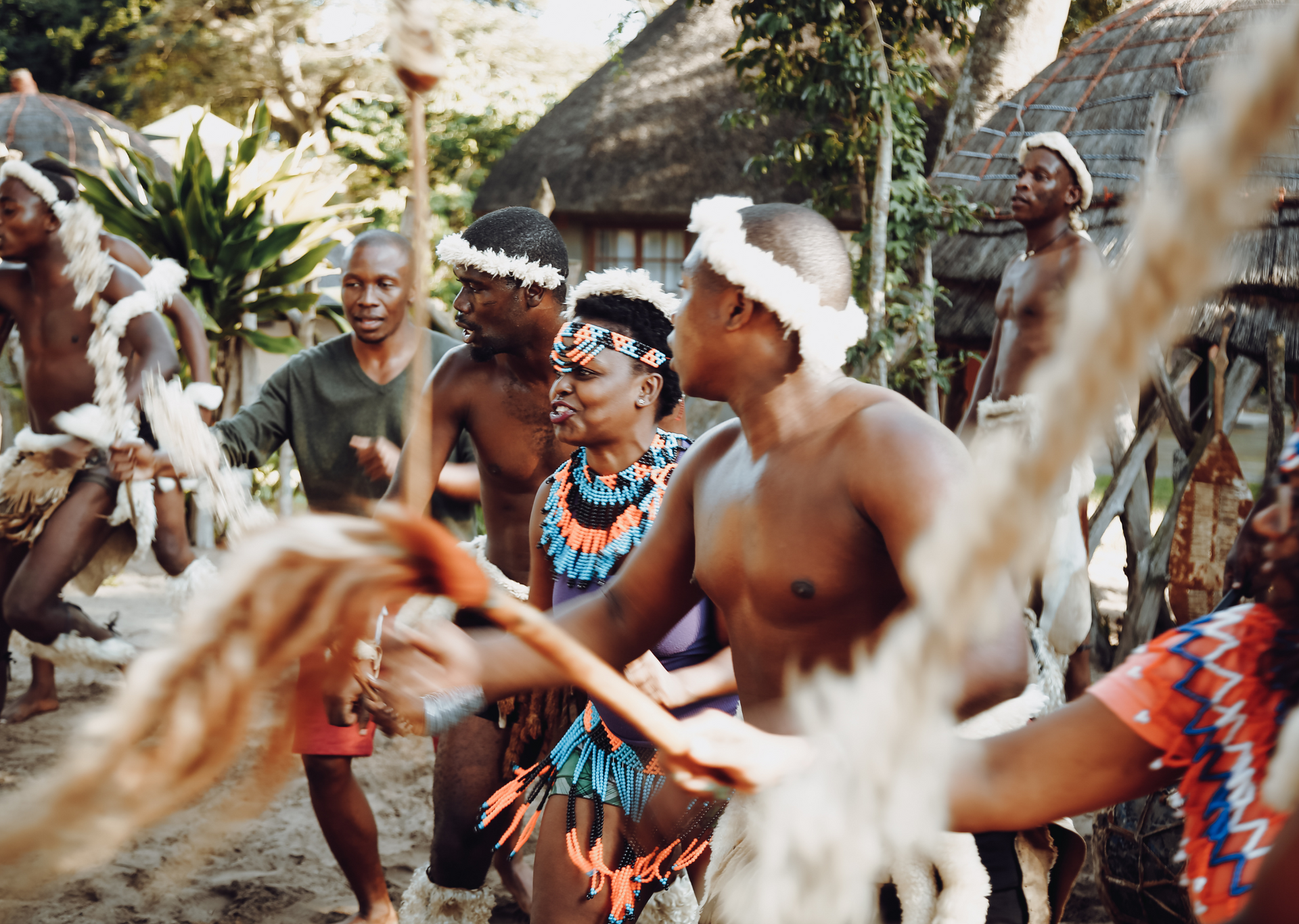 People in traditional African attire dancing in front of thatched-roof huts. They wear headbands, beaded necklaces, and animal hide skirts.