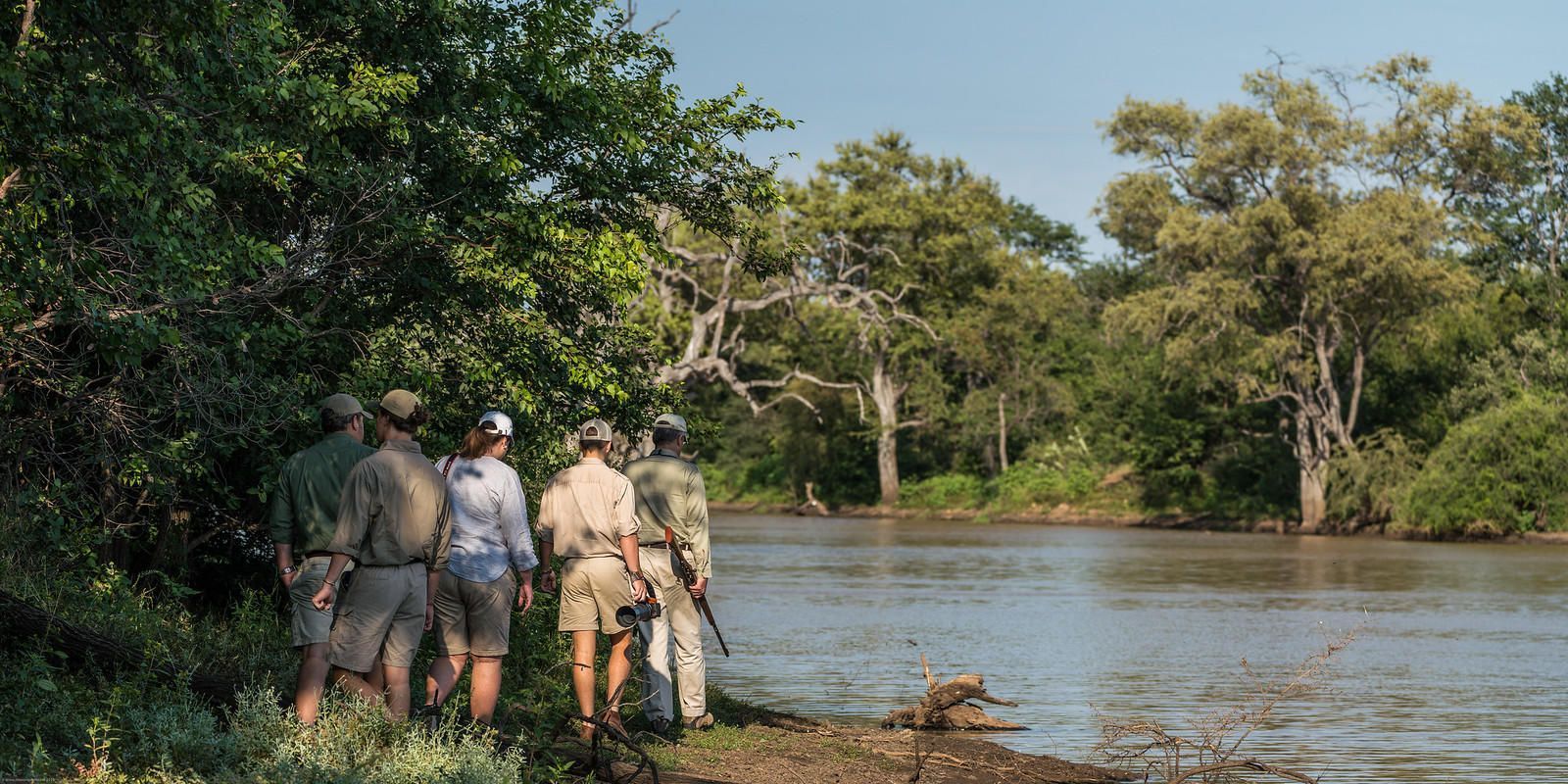 A group of people standing near a river, surrounded by trees.