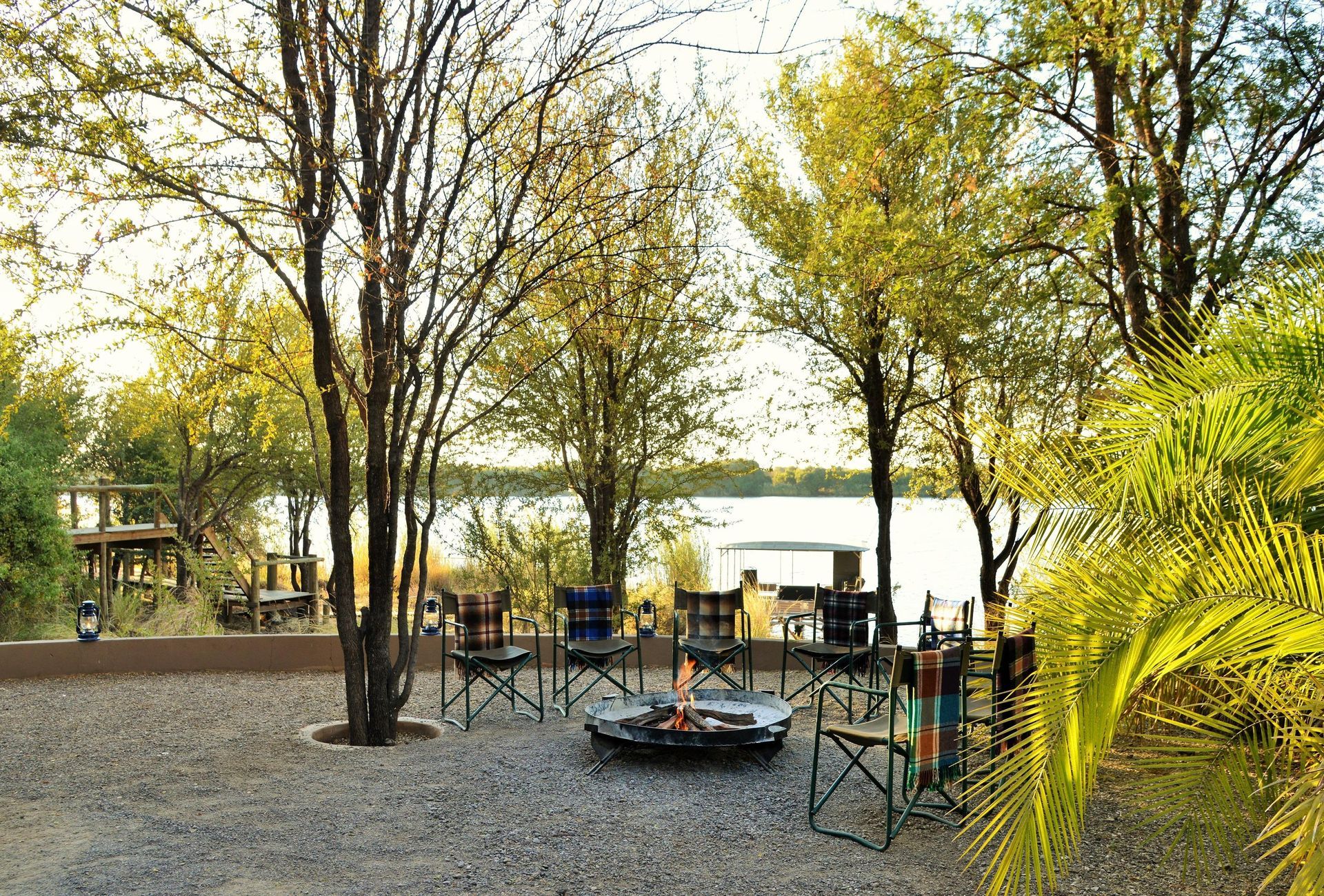Camp chairs circle a fire pit on a gravel patio, with a river and trees in the background.