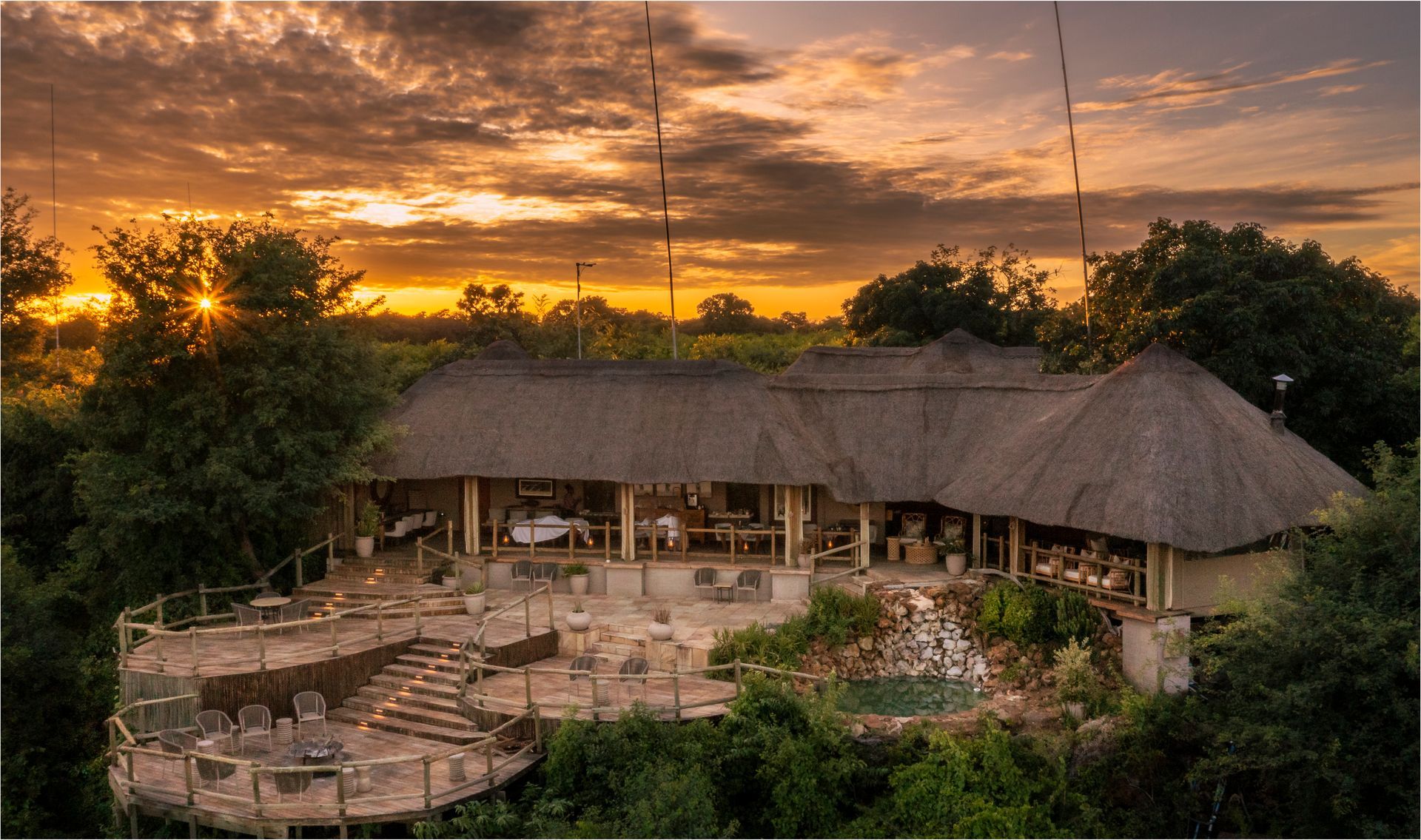 African lodge with thatched roof at sunset, overlooking a lush landscape.