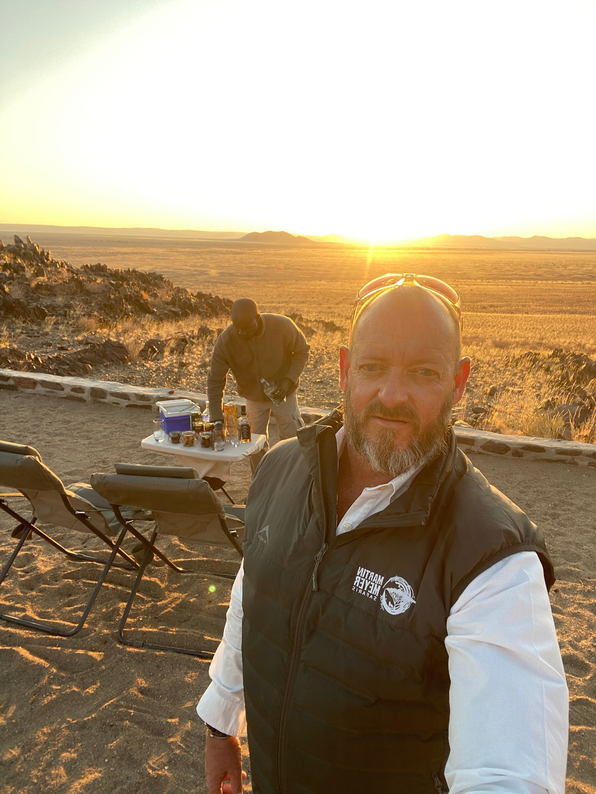 A man with a beard is standing in front of a sunset in the desert. Namibia Safari