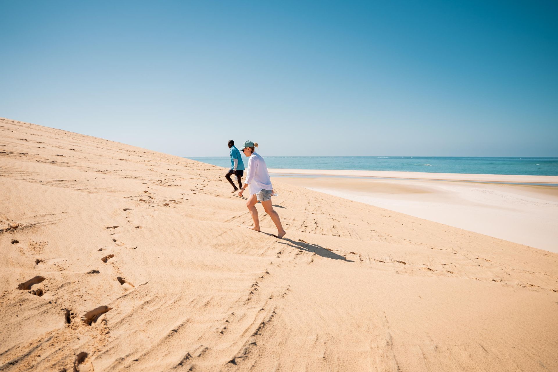 Two people walking on a sandy beach with the ocean in the background under a blue sky.