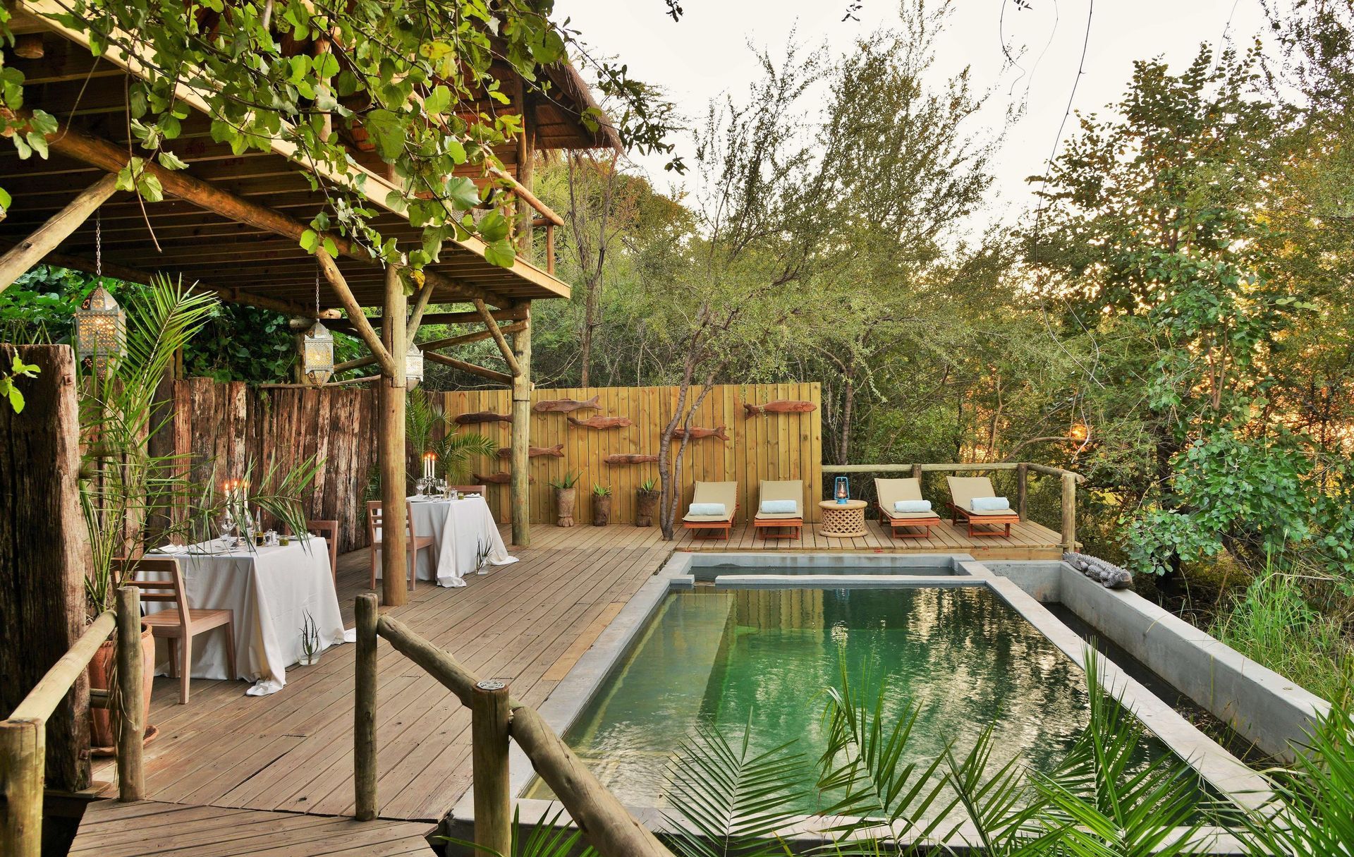 A large swimming pool is surrounded by a wooden deck with tables and chairs.