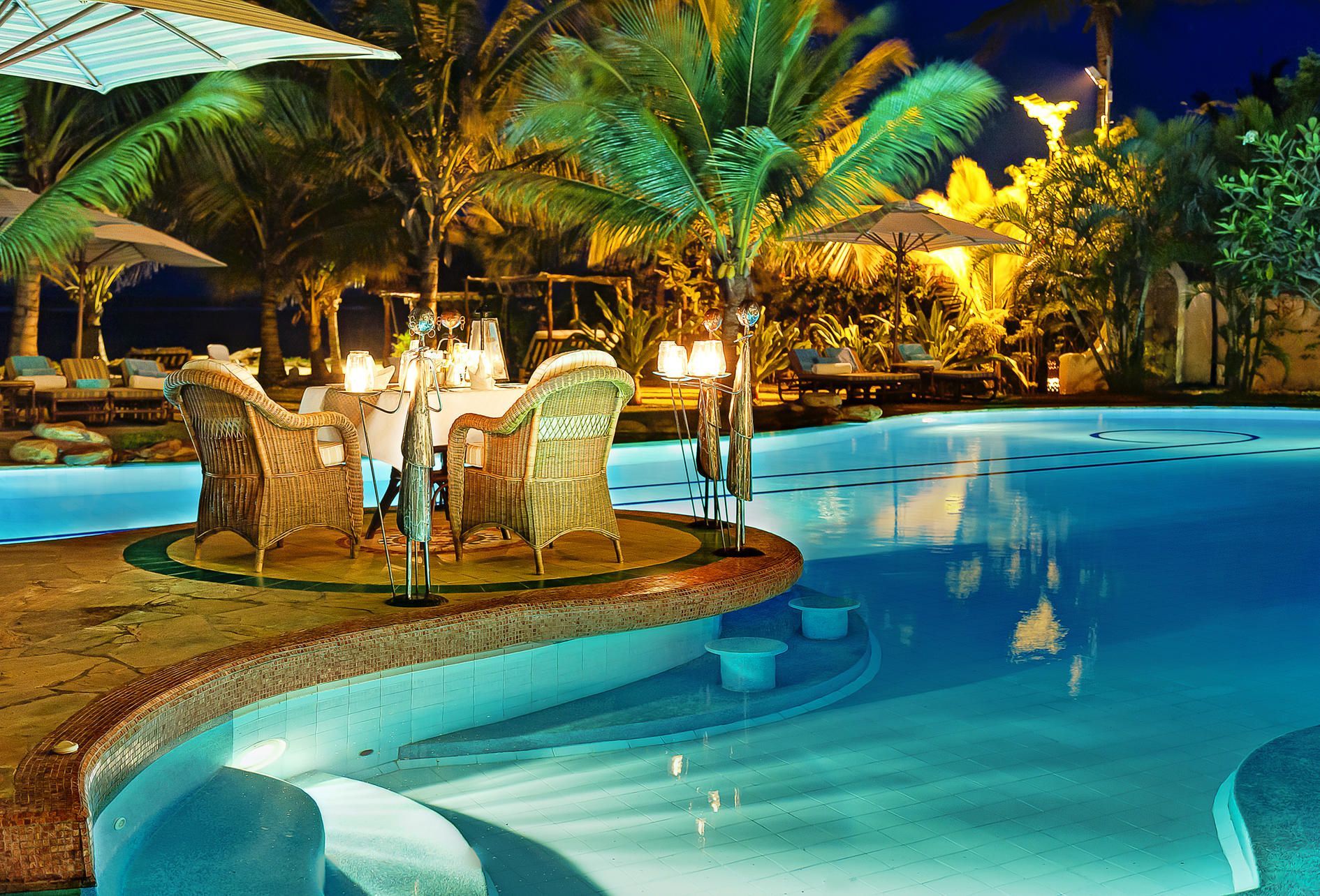 A resort pool and beach scene at sunset with umbrellas, palm trees, and ocean views.