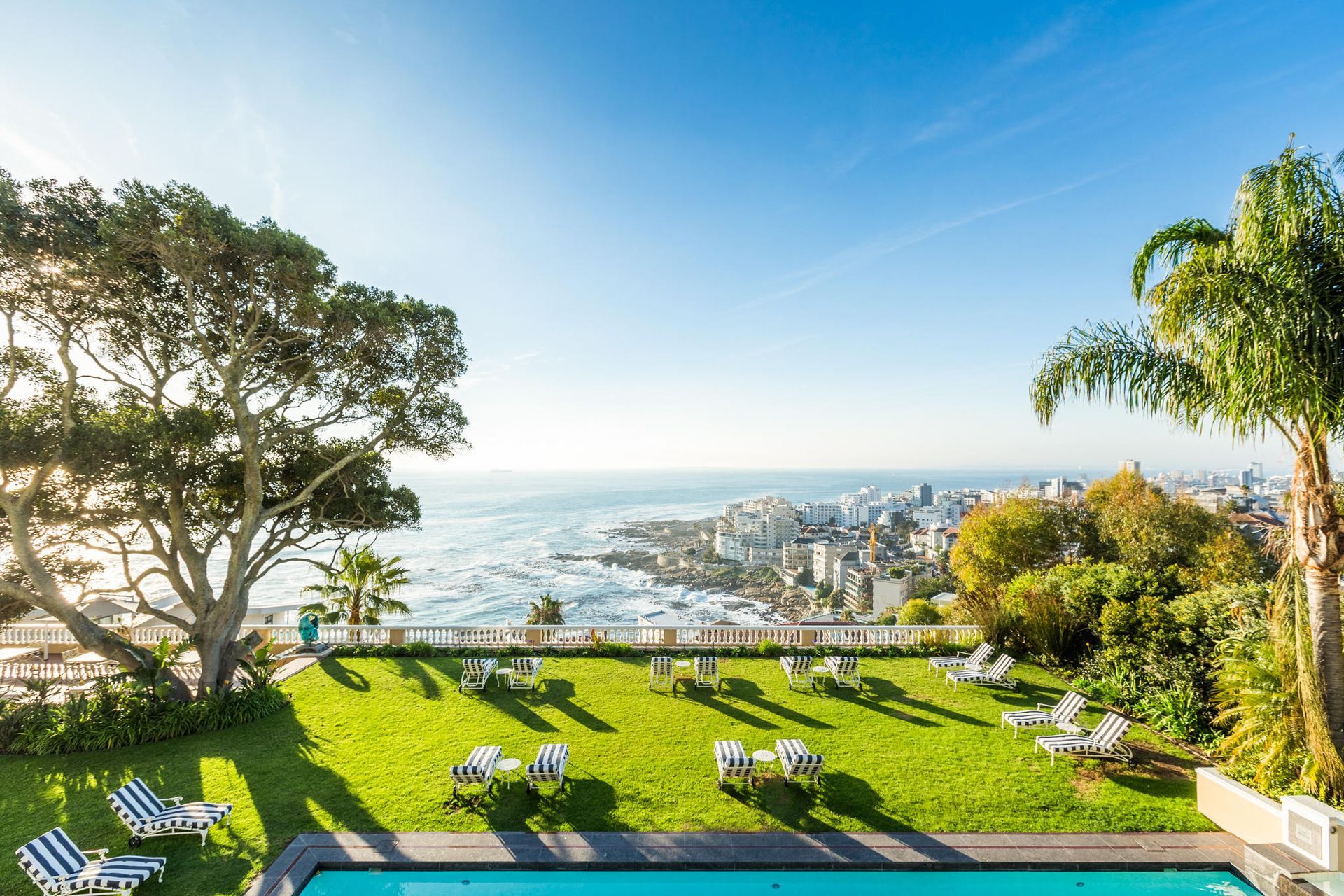Green lawn with lounge chairs, pool, ocean view, and city under blue sky.