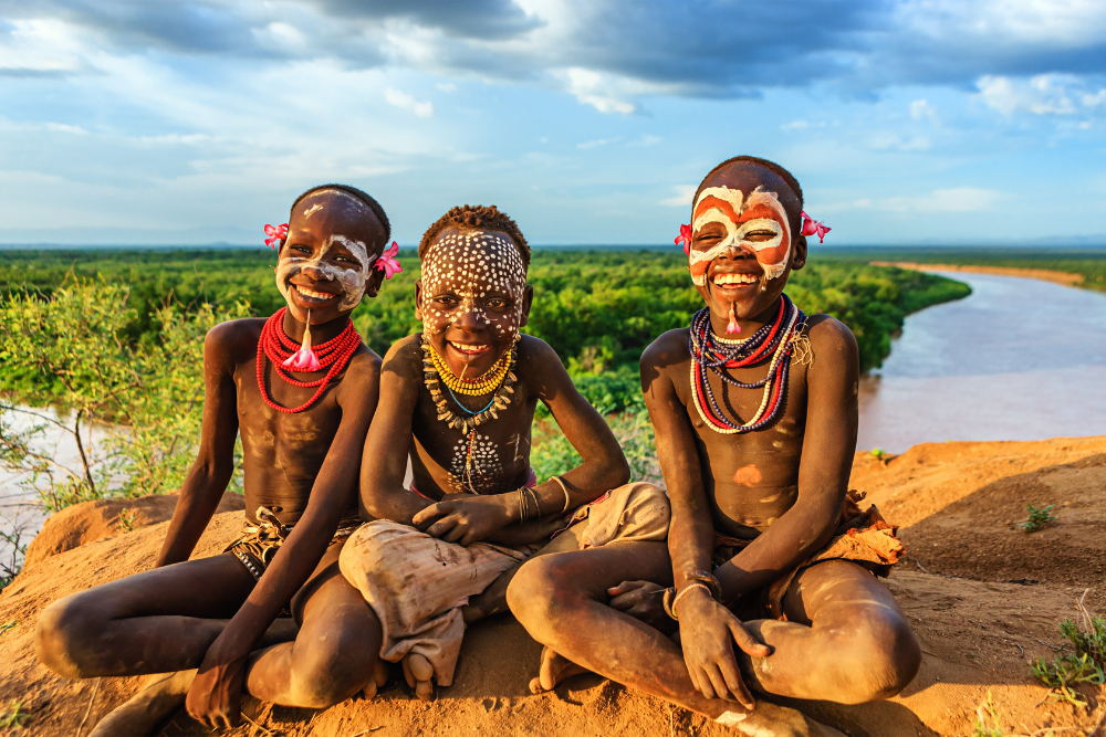 Three young boys with face paint are sitting on a rock near a river.