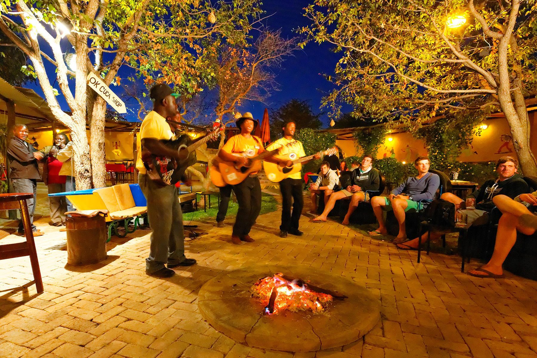 Band playing around a fire pit at night; people watching, trees and lights overhead.