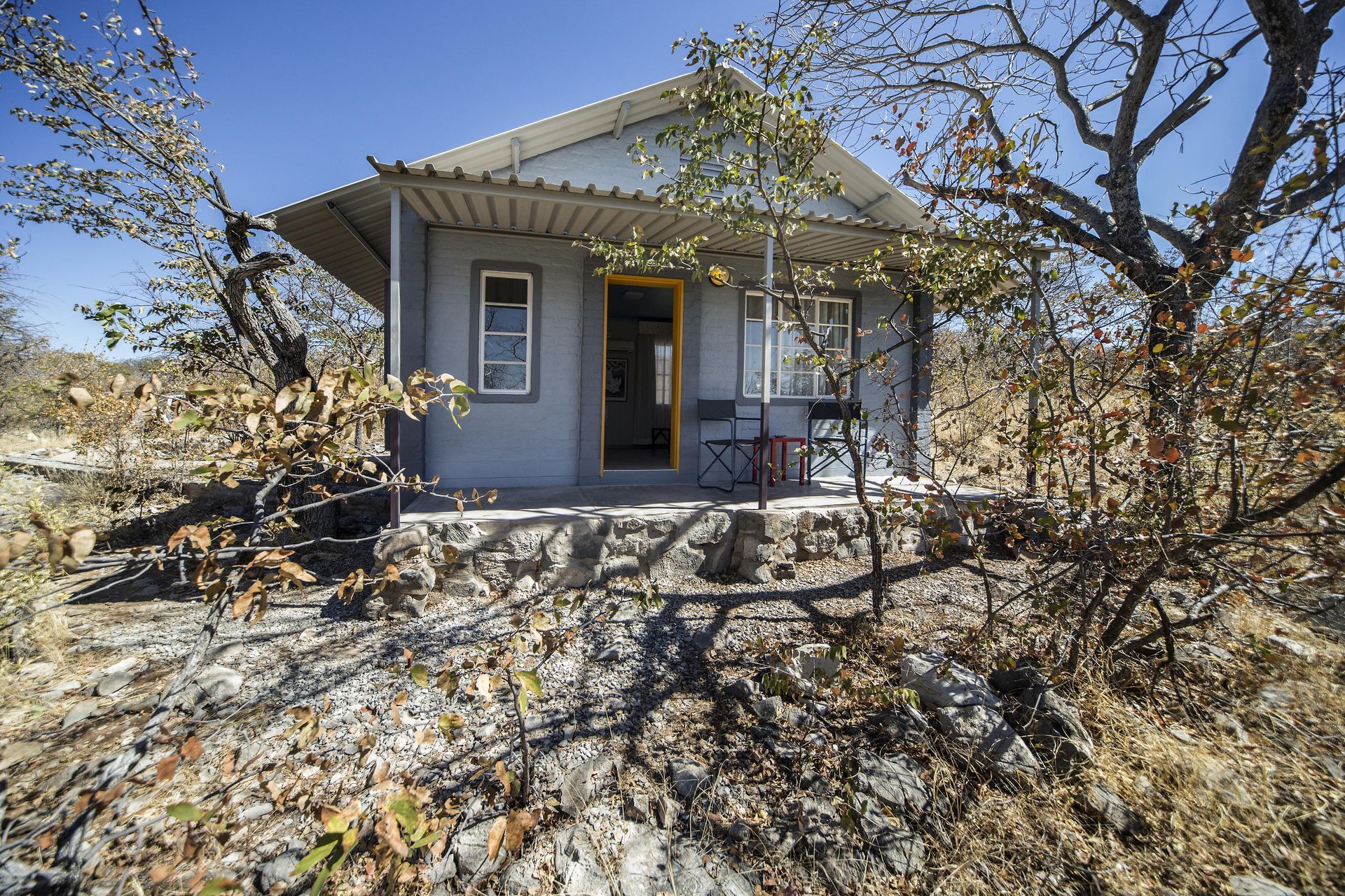 Small, gray cottage with yellow door and chairs on a stone patio, in a dry, sunny landscape.
